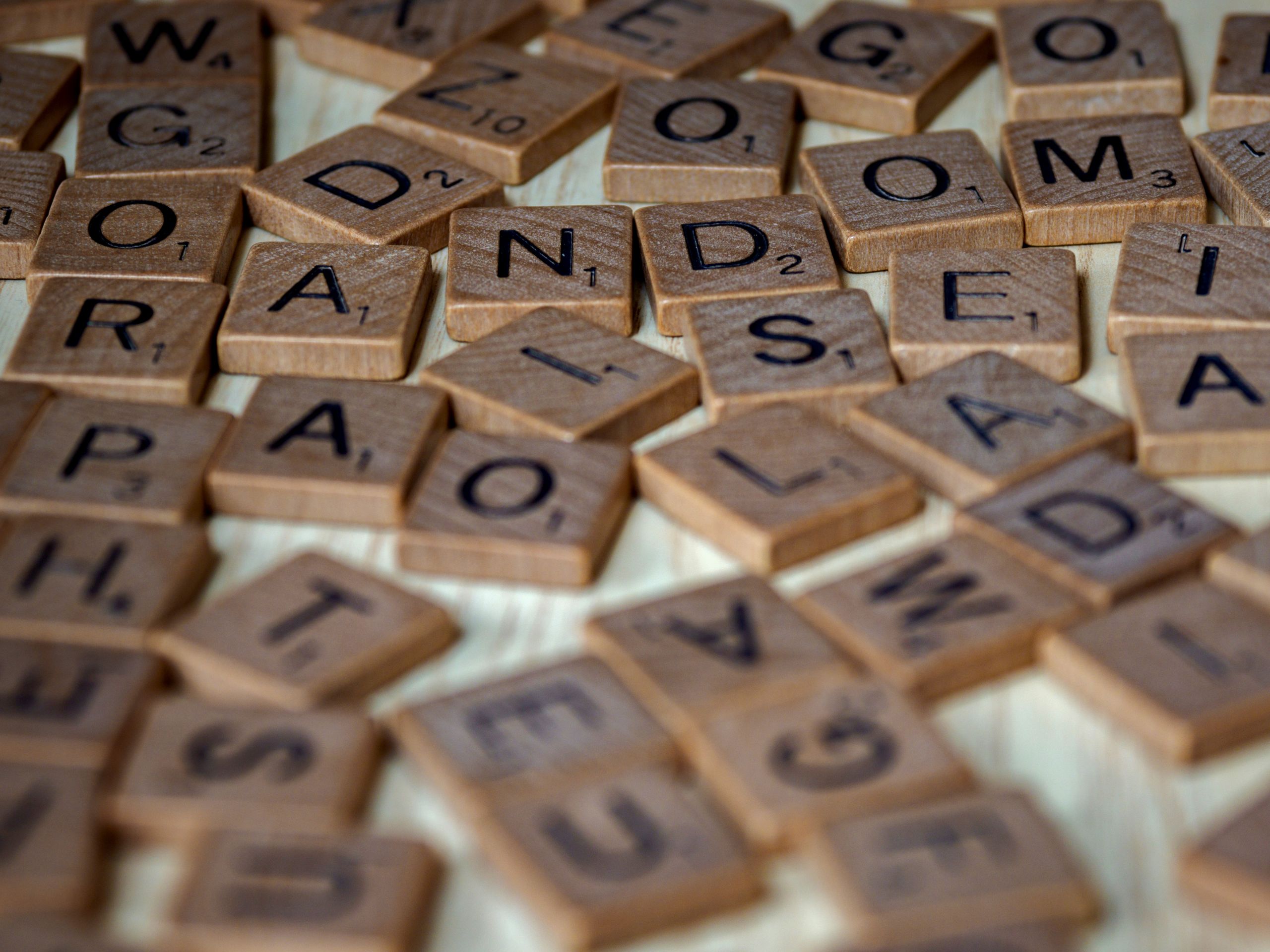 Le lexique de la com’ : le jargon traduit en vrai français A close-up photo showing scattered wooden Scrabble tiles with letters and point values.